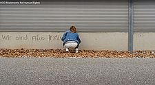 Videostill einer Schülerin von hinten, die mit Wasser einen Satz auf eine Wand im öffentlichen Raum sprüht.