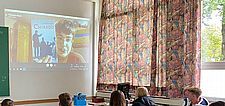 Foto von einem Klassenraum mit einigen Schüler/innen, an der Wand eine Projektion der Skype-Kameraansicht der Autorin Theodora Bauer mit ihrem Buch
