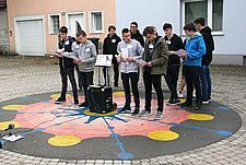 Foto von 12 Schülern, die mit Textheften und zwei Mikrofonen im öffentlichen Raum performen