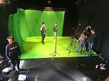 Foto von zwei Schüler/innen in einem grün ausgekleideten Studioeck, andere bedienen Scheinwerfer und Kamera